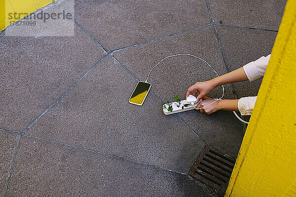 Hände einer jungen Frau  die ihr Mobiltelefon über eine Steckdose auflädt  und Pflanzen auf dem Boden