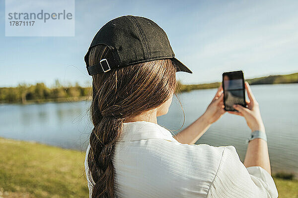 Junge Frau mit Mütze fotografiert See auf Smartphone