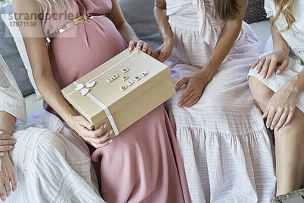 Hände einer schwangeren Frau  die zu Hause mit Freunden sitzt und eine Geschenkbox öffnet