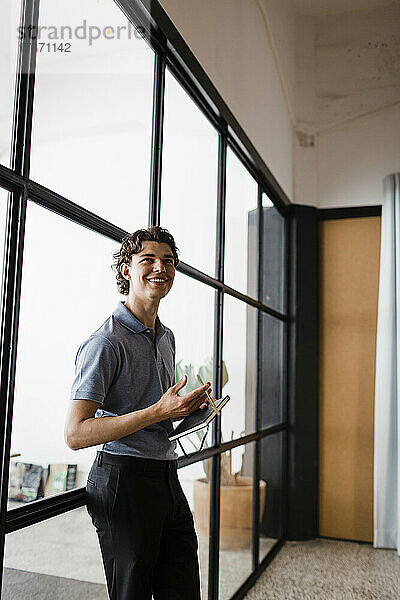 Happy young businessman standing with book leaning on glass wall