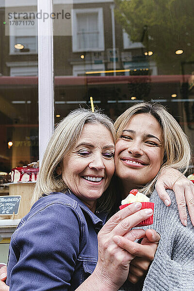 Lächelnde Mutter und Tochter mit Cupcake umarmen sich vor dem Laden