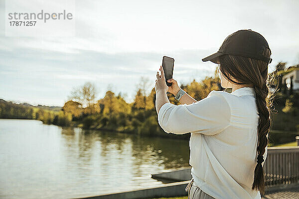 Frau mit Mütze fotografiert den See an einem sonnigen Tag per Smartphone