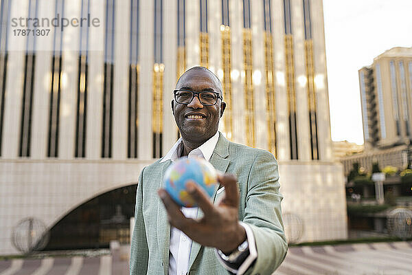 Glücklicher reifer Geschäftsmann mit Globusfigur  der vor dem Gebäude steht