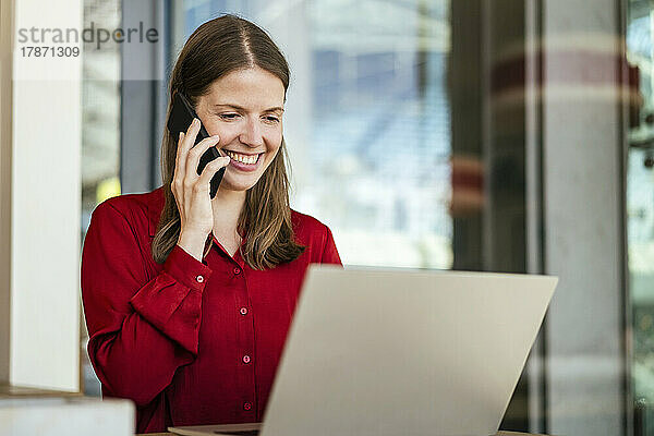 Lächelnde Geschäftsfrau  die im Büro mit einem Smartphone spricht und einen Laptop benutzt