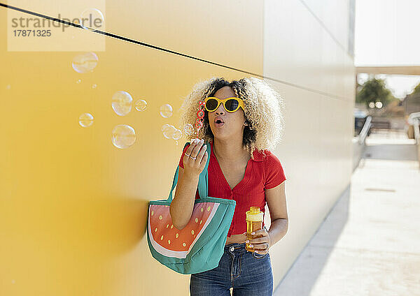 Junge Frau mit Sonnenbrille bläst Seifenblasen an der gelben Wand