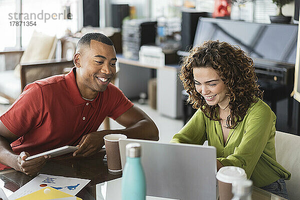Lächelnde junge Geschäftsfrau teilt Laptop mit Kollegin  die am Schreibtisch im Büro sitzt
