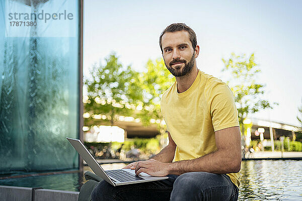 Lächelnder junger Freiberufler mit Laptop sitzt vor dem Teich