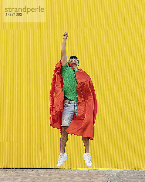 Junger Mann mit rotem Umhang springt vor gelbe Wand