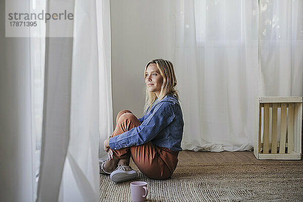 Frau sitzt zu Hause auf einem Teppich und umarmt Knie am Fenster