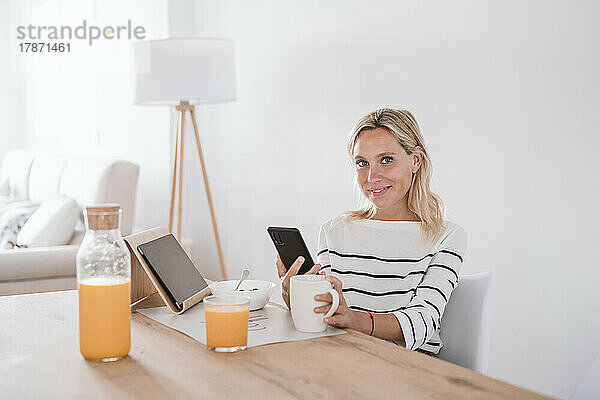 Lächelnde Frau mit Kaffeetasse und Mobiltelefon zu Hause