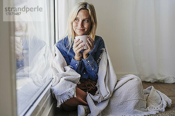Junge Frau  in eine Decke gehüllt  hält eine Kaffeetasse in der Hand und sitzt zu Hause am Fenster
