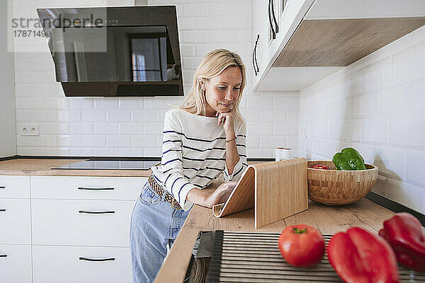 Junge Frau mit Hand am Kinn schaut sich Rezept auf Tablet-PC in der heimischen Küche an