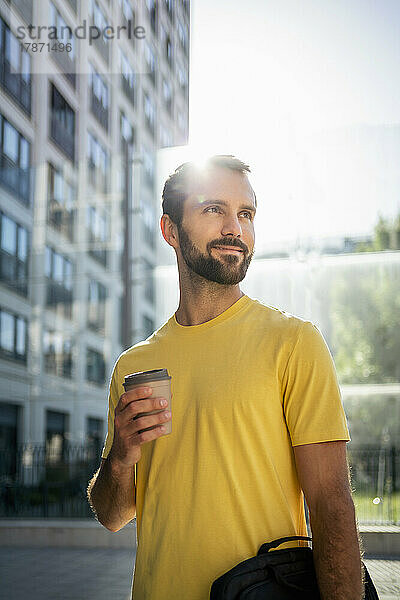 Lächelnder junger Geschäftsmann mit Kaffeetasse  der vor dem Gebäude steht