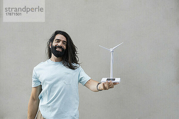 Lächelnder Hipster-Mann mit langen schwarzen Haaren  der ein Windturbinenmodell vor der Wand hält