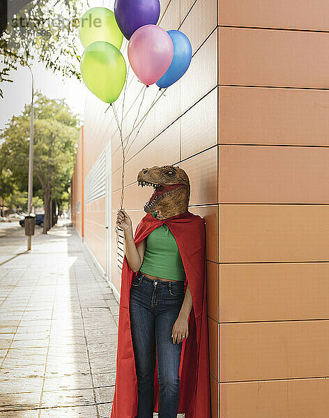 Junge Frau mit Krokodilmaske steht mit bunten Luftballons an der Wand