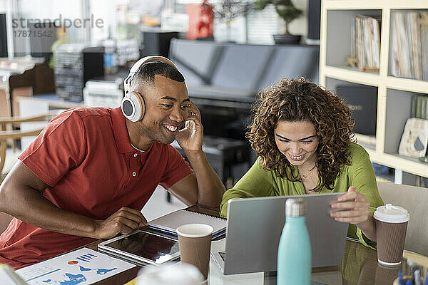Lächelnde Geschäftsfrau teilt Laptop mit Kollege  der kabellose Kopfhörer im Büro trägt