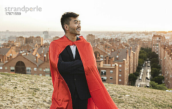 Glücklicher junger Geschäftsmann mit rotem Superheldenumhang  der auf einem Hügel steht