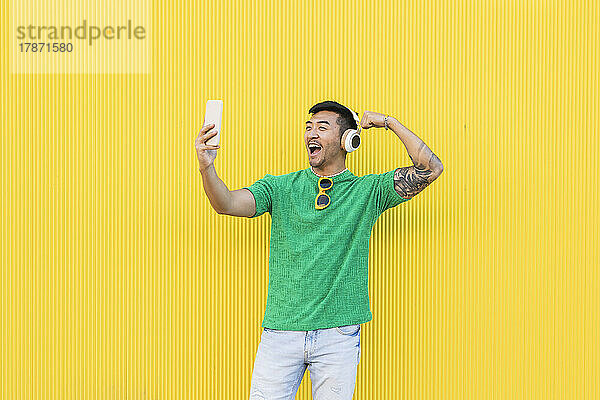Glücklicher junger Mann zeigt Bizeps bei Videoanruf über Smartphone vor gelber Wand