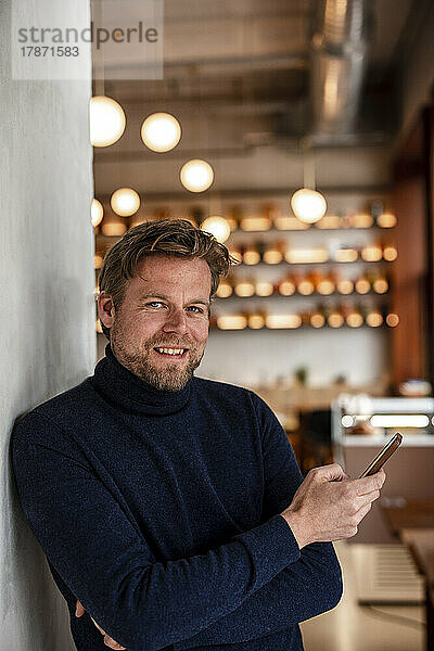Lächelnder Geschäftsmann mit Mobiltelefon lehnt an der Wand im Café