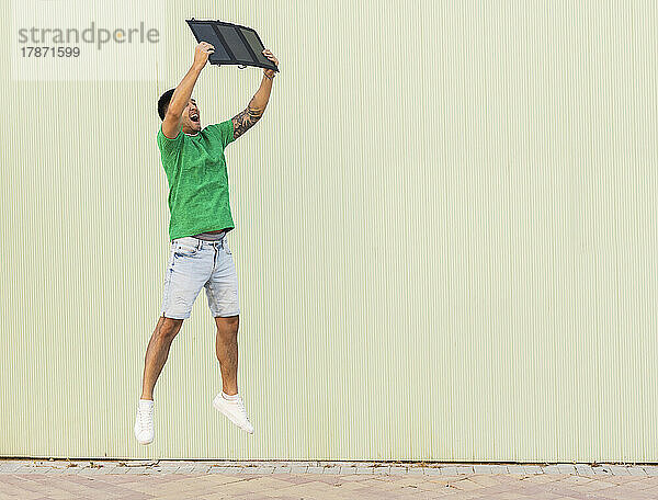 Glücklicher junger Mann springt mit Solarpanel vor der Wand