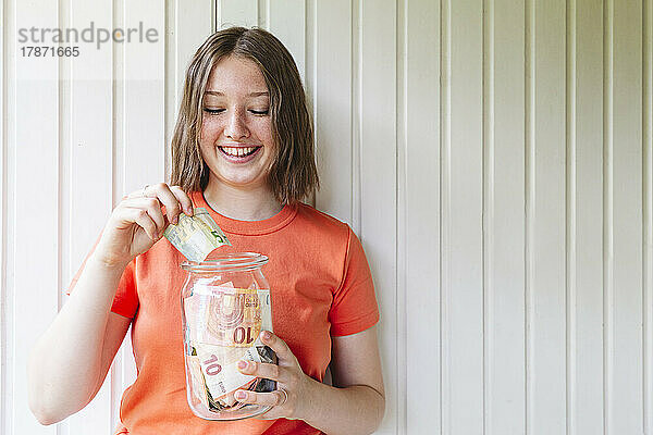 Lächelndes Mädchen hält Glasgefäß mit europäischer Währung vor der Wand