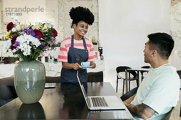 Lächelnde Kellnerin serviert Kaffee an Kunden  die mit Laptop im Café sitzen