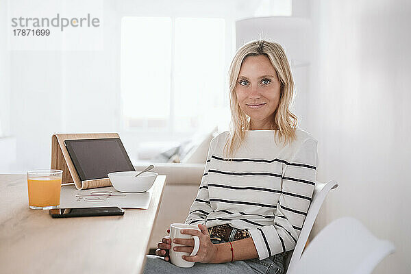 Lächelnde Frau mit Kaffeetasse und Tablet-PC auf dem heimischen Tisch
