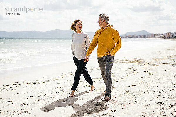 Lächelnde Frau und Mann halten sich an den Händen und gehen am Strand entlang