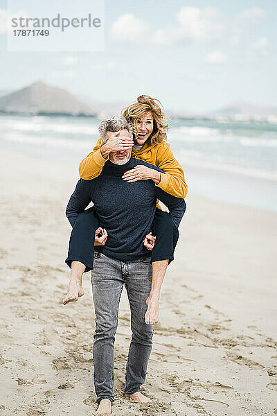 Fröhliche Frau bedeckt die Augen eines Mannes mit der Hand und genießt die Huckepackfahrt am Strand