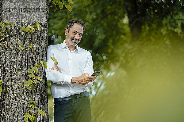 Lächelnder reifer Geschäftsmann mit Smartphone am Baum