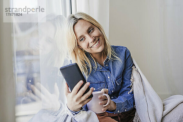 Lächelnde Frau benutzt Smartphone und lehnt sich zu Hause an Fensterglas