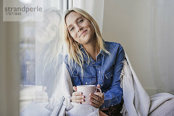 Lächelnde Frau mit Kaffeetasse lehnt zu Hause an Glasfenster