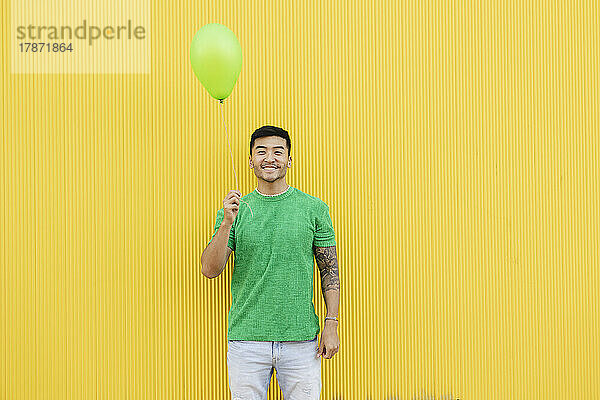 Smiling young man holding green balloon in front of wall