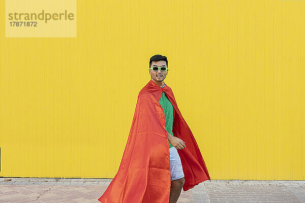 Young man with cape and sunglasses standing in front of yellow wall