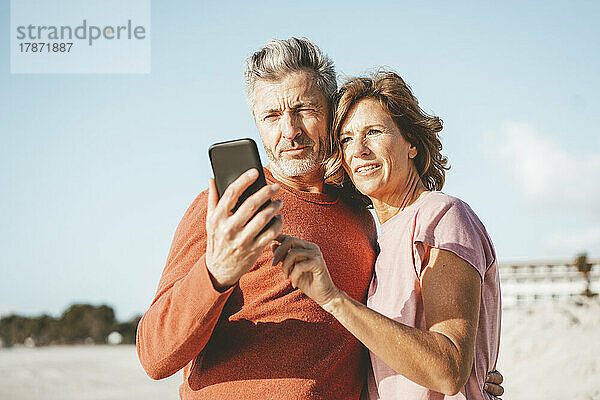 Lächelndes älteres Paar  das am Strand sein Mobiltelefon benutzt