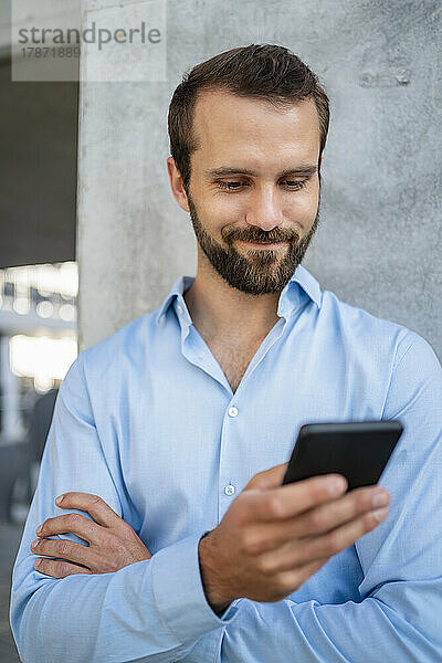 Lächelnder junger Geschäftsmann mit Smartphone vor der Wand