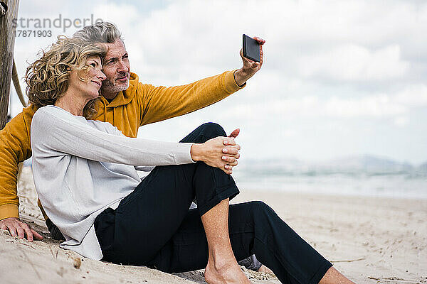 Smiling man taking selfie with woman through mobile phone at beach