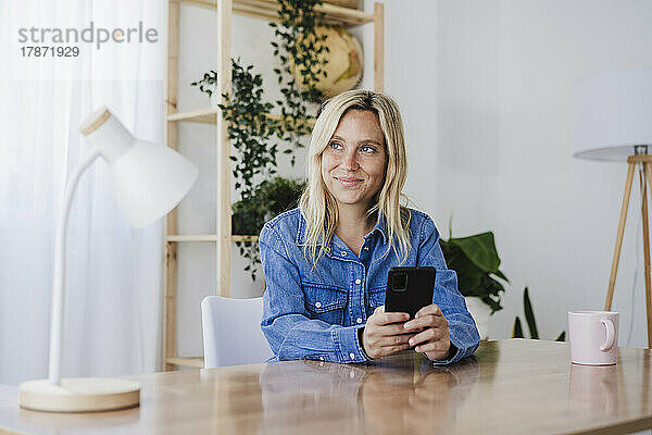 Nachdenklicher Freiberufler mit Smartphone sitzt am Schreibtisch im Homeoffice