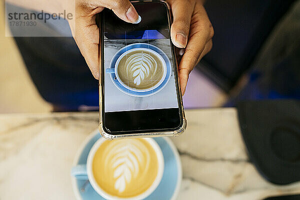 Hände einer jungen Frau  die im Café eine Kaffeetasse über ihr Smartphone fotografiert