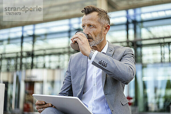 Geschäftsmann trinkt Kaffee und hält ein digitales Tablet vor dem Gebäude