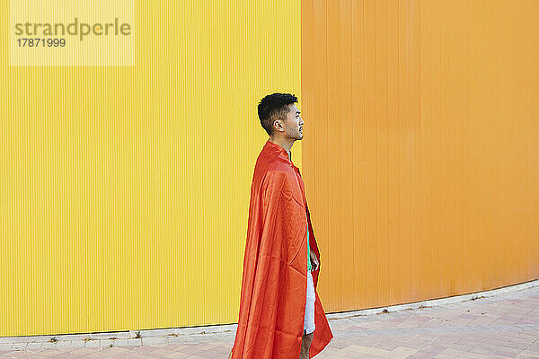 Junger Mann mit rotem Superheldenumhang steht vor der Wand