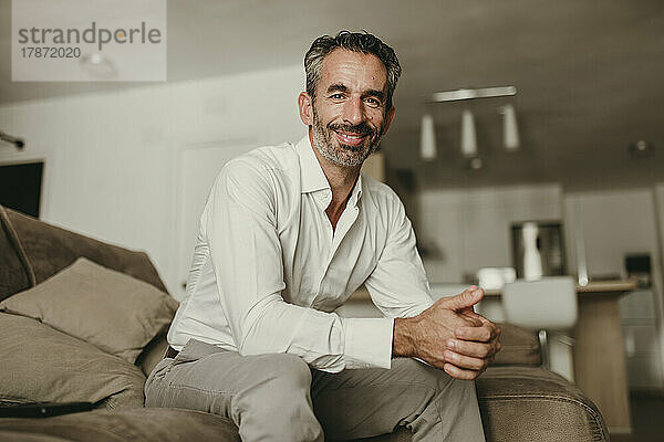 Lächelnder Geschäftsmann sitzt mit gefalteten Händen auf dem Sofa zu Hause