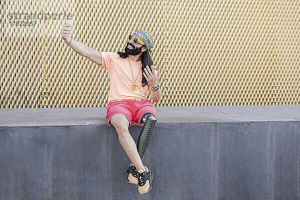 Glücklicher Mann mit Beinprothese  der ein Selfie mit dem an der Wand sitzenden Smartphone macht