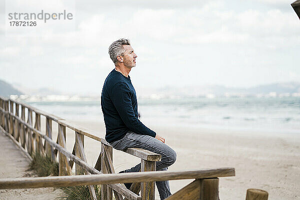 Nachdenklicher reifer Mann sitzt auf dem Geländer am Strand