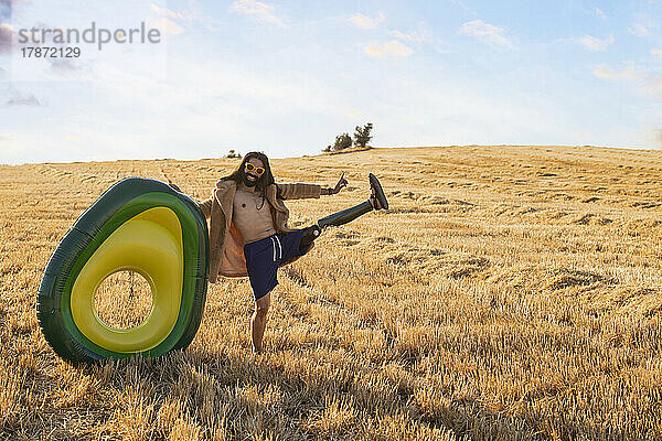 Glücklicher Hipster-Mann mit Beinprothese  der an einem sonnigen Tag mit einem aufblasbaren Ring auf dem Feld balanciert