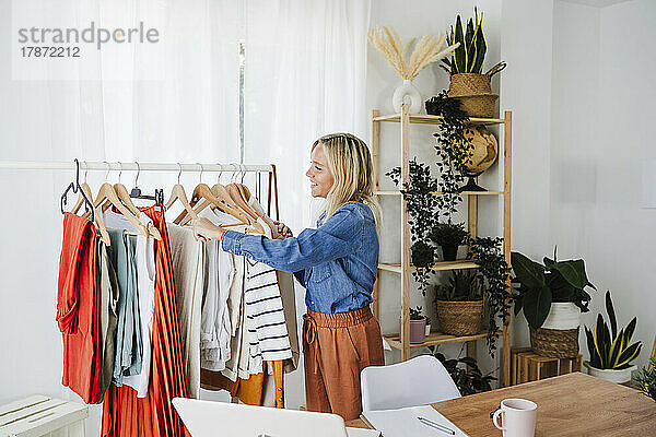 Lächelnder Modedesigner hängt Kleidung am Kleiderständer im Heimbüro