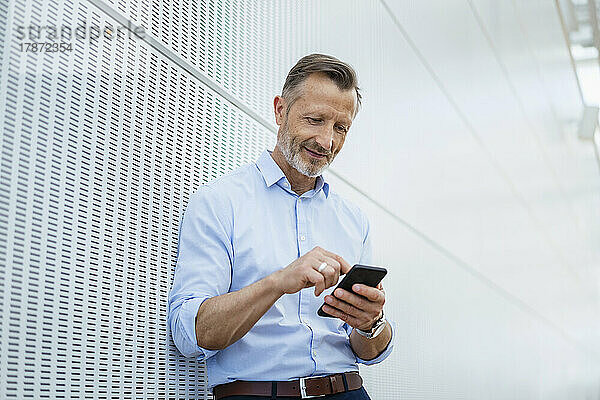 Lächelnder Geschäftsmann  der sein Mobiltelefon an der Wand benutzt