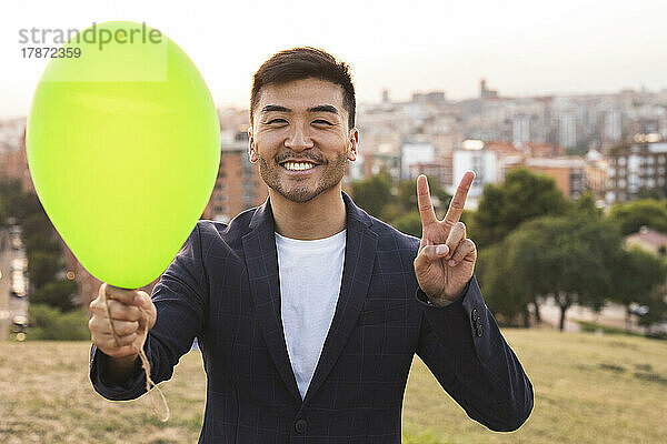Fröhlicher junger Geschäftsmann mit grünem Ballon  der bei Sonnenuntergang ein Friedenszeichen zeigt