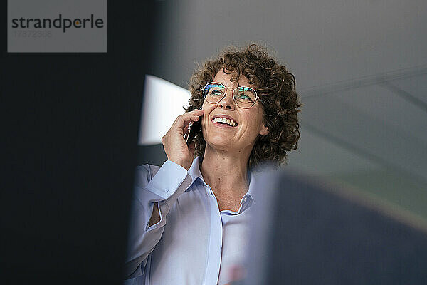 Glückliche Geschäftsfrau mit Brille  die im Büro auf dem Smartphone spricht