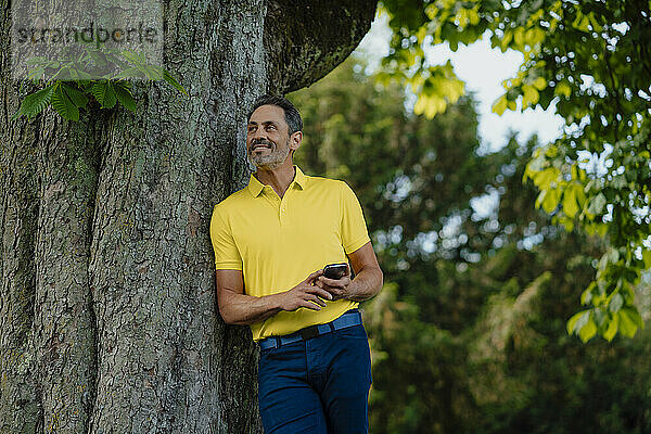 Lächelnder Mann hält Smartphone in der Hand und lehnt an Baumstamm